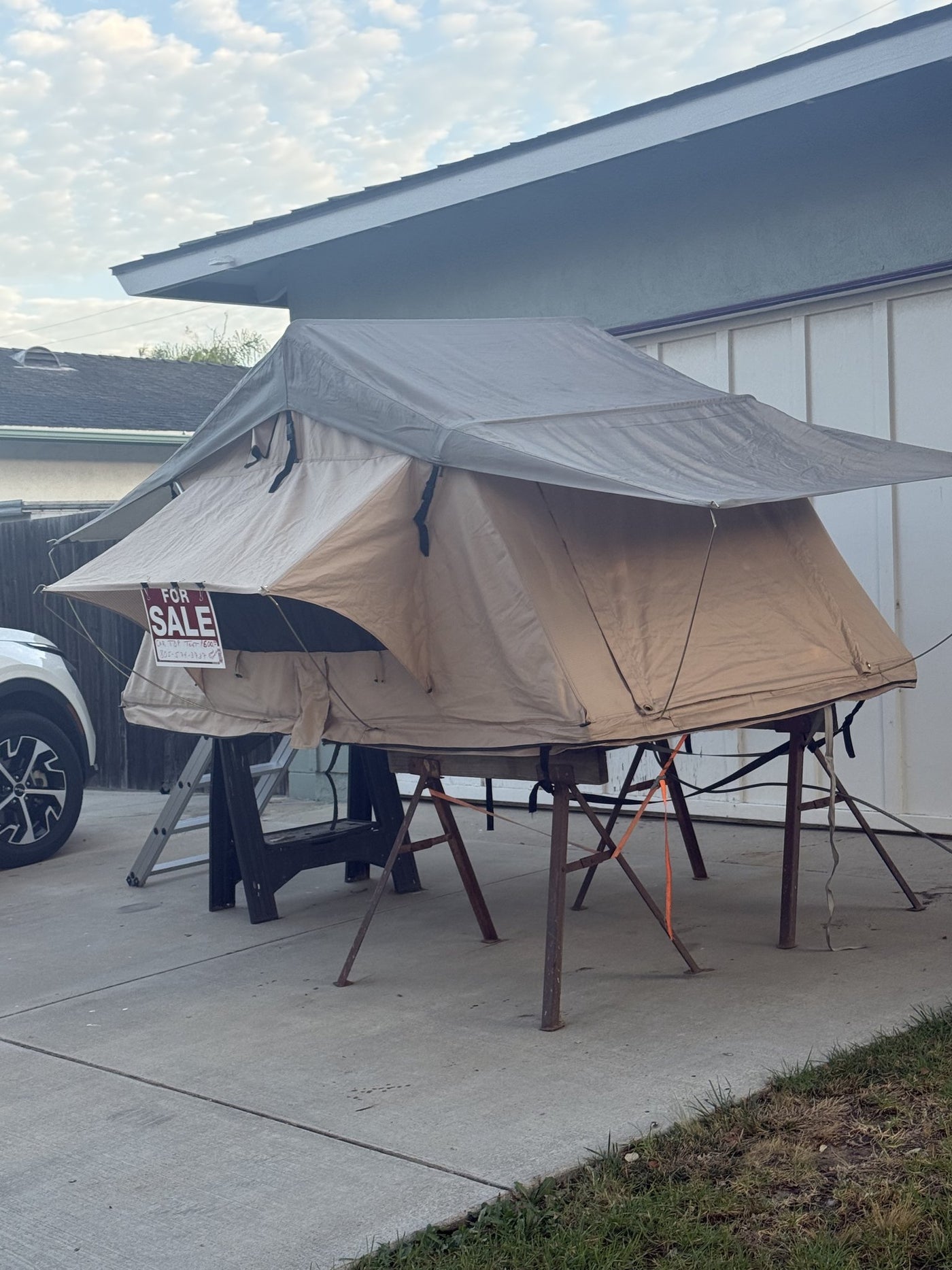 The tent standing open on sawhorses in a driveway with a FOR SALE sign clipped to it, showing current condition of the tan canopy and rainfly — photo 3