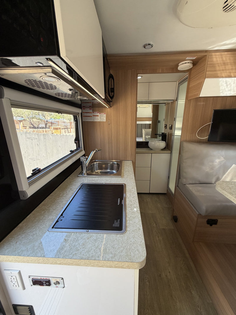 Galley looking aft down the trailer: stone-look countertop with stainless sink and covered Dometic cooktop, overhead microwave, wood-grain cabinetry, vinyl plank flooring and the bathroom and bedroom at the far end — photo 7