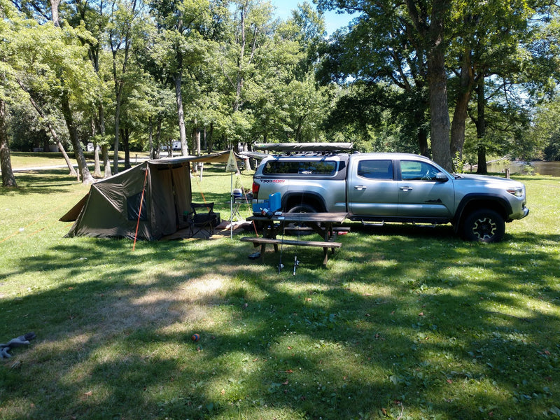 Oztent RV-4 canvas touring tent pitched at a sunny wooded campsite with the front awning extended on poles, set up alongside a silver Toyota Tacoma with a bed canopy and rooftop tent — photo 1