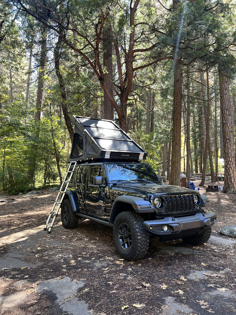 The BDV Duo opened on the Jeep's roof rack at a pine-forest campsite, telescoping aluminum ladder extended down to the ground, seen from the front three-quarter — photo 2