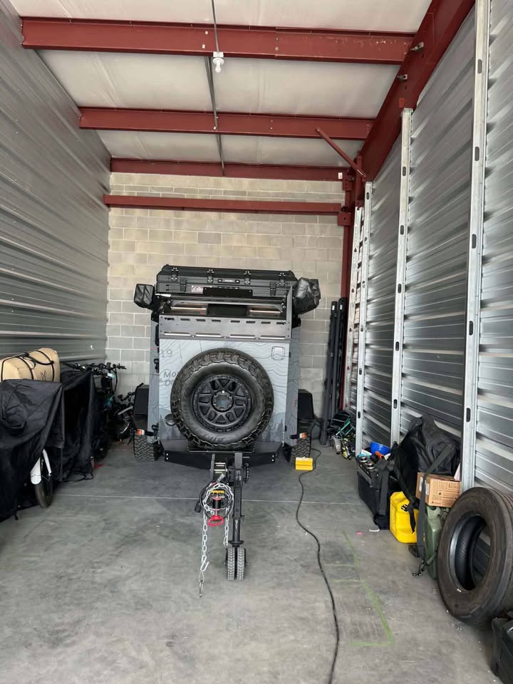 Front view of the XPedition Voyager Sky XE parked nose-in inside an enclosed storage unit, showing the spare tire on the front rack, roof cargo tray, topographic-map body graphics, tongue jack and safety chains — photo 5