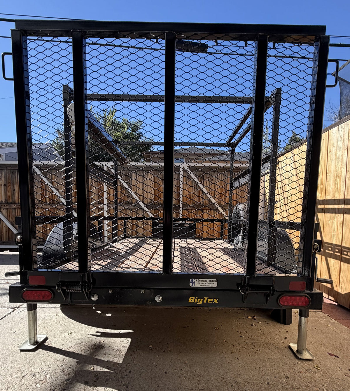 Straight rear view with the expanded metal mesh drop gate closed and both rear stabilizing jacks lowered onto the concrete, showing the Big Tex badge, red LED tail lights and the overhead rack running the length of the deck — photo 4