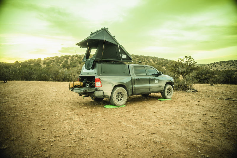 NGHTOWL Offroad aluminum camper shell mounted on a 2019+ Ram 1500 short bed with the rooftop tent fully deployed, rear three-quarter view on open desert ground, the shell's rear hatch propped open over a lowered tailgate with gear loaded and green traction boards under the rear tires — photo 1