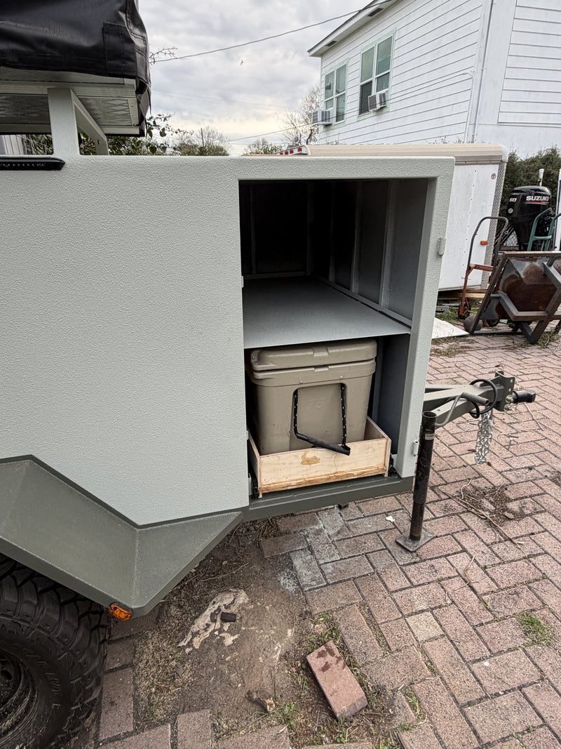 Side hatch open on the passenger side showing the upper shelf and the lower slide-out drawer with a tan 75-quart ice chest strapped in place, trailer tongue and jack visible at right — photo 5