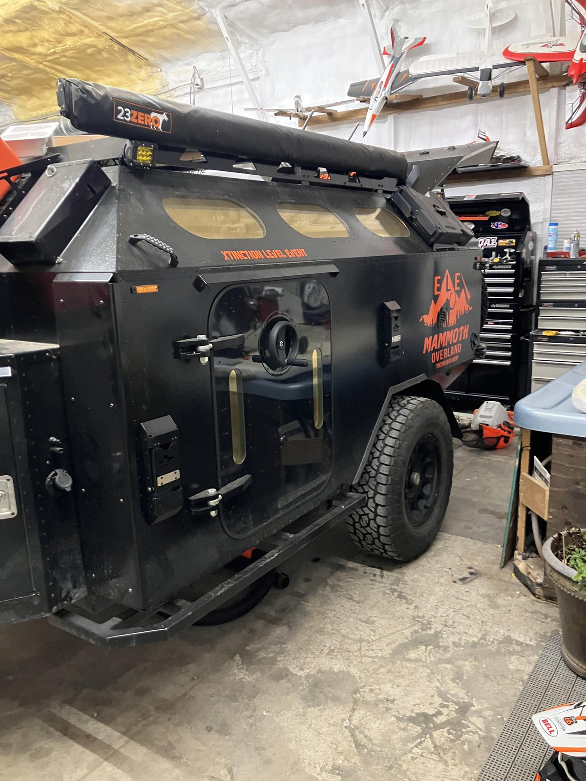 Blacked-out 2023 Mammoth Overland ELE off-road trailer parked in a shop, shot from the rear three-quarter, showing the MAMMOTH OVERLAND and XTINCTION LEVEL EVENT graphics on the side panel, the rolled 23ZERO awning on the roof rack, the round-porthole cabin windows and the side entry door — photo 1