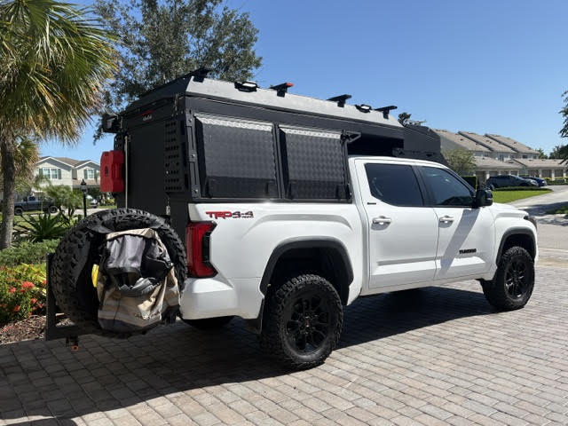 Rear three-quarter view on a paver driveway with the camper closed, showing the checker-plate rear panel, red Rotopax fuel cell mounted on the side and the covered spare tire on the swing-arm carrier — photo 4