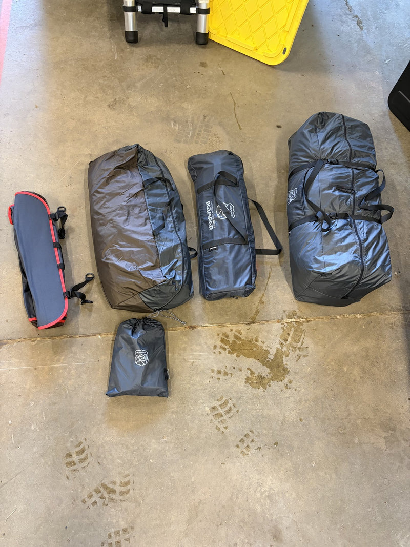 The included accessory bags laid out on a garage floor, iKamper branded storage duffels holding the annex, awning and canopy pieces plus a smaller zip pouch and a rolled grey and red bag — photo 9