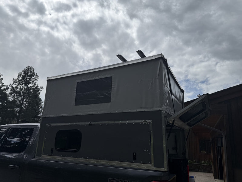 Three-quarter rear view of the OVRLND Bivy camper on a dark pickup with the roof popped up and the rear flip-up door raised on its struts, showing the riveted aluminum construction and roof rack rails — photo 2