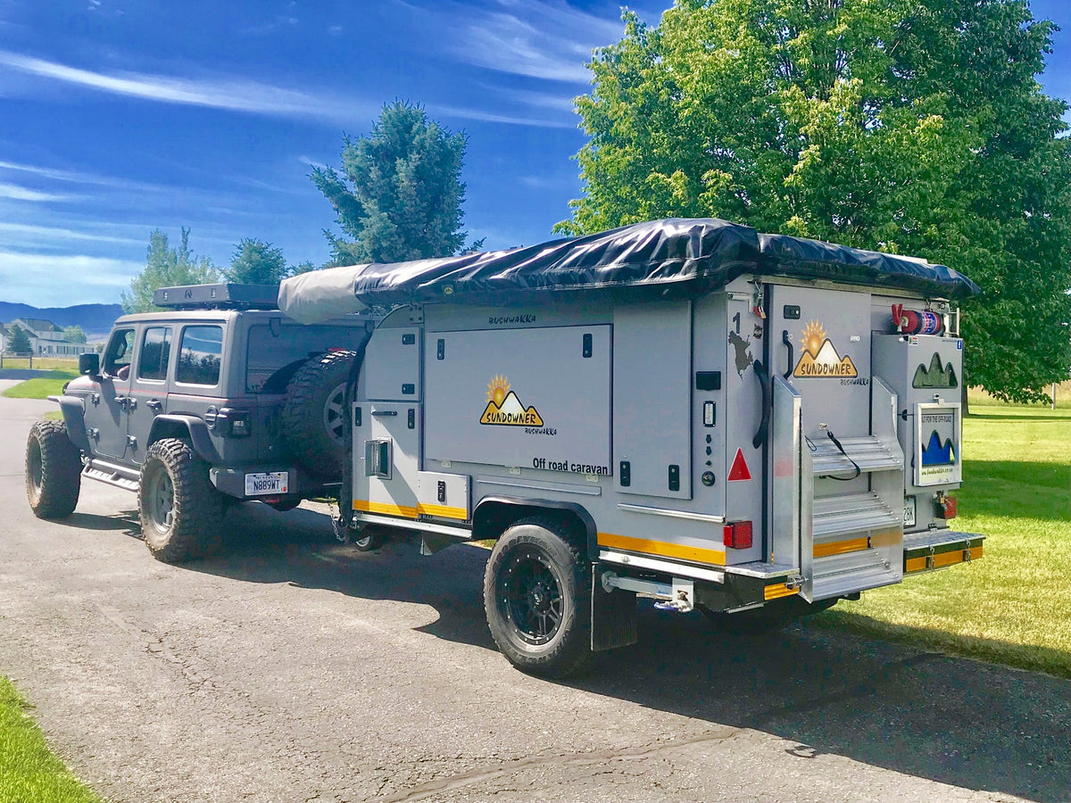 2019 Bushwakka Sundowner off-road caravan in travel mode, hitched to a grey Jeep Wrangler on a paved drive, showing the white body with Sundowner sun-and-mountain graphics, yellow and black hazard striping, side storage hatches, rolled awning on the roof and black alloy wheels on all-terrain tires