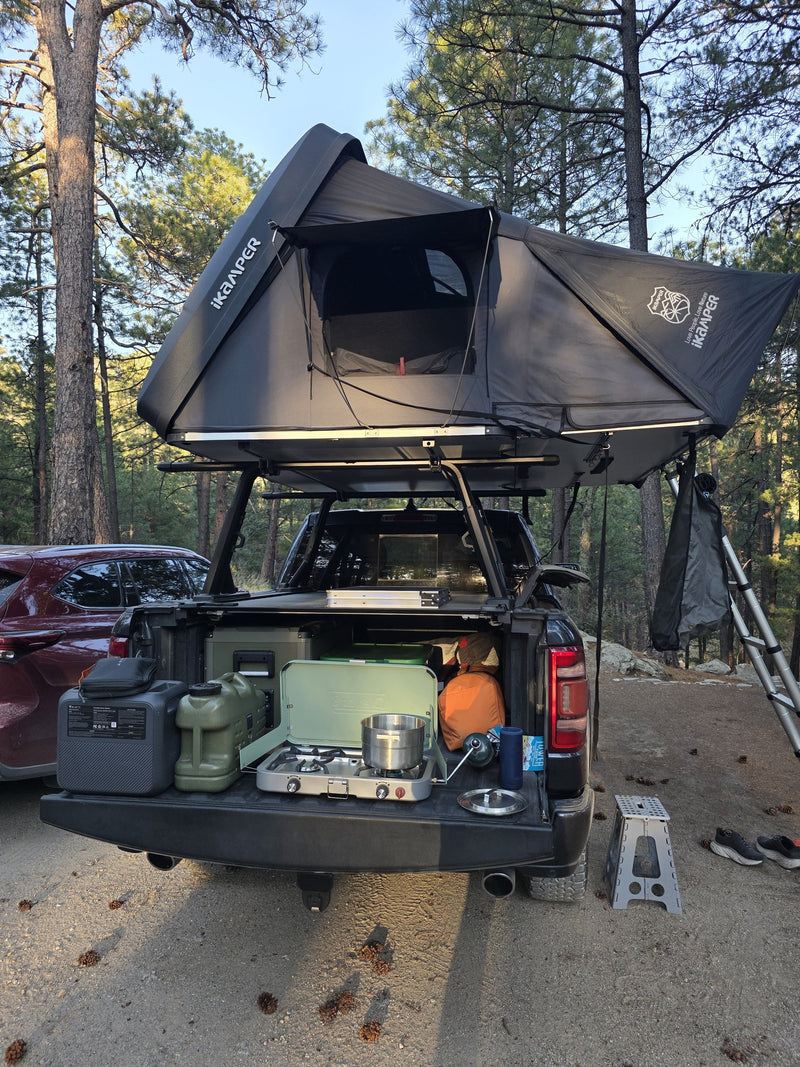 iKamper DLX rooftop tent fully open on a Ram 1500 at a pine-forest campsite, tailgate down with the camp kitchen set up — photo 1