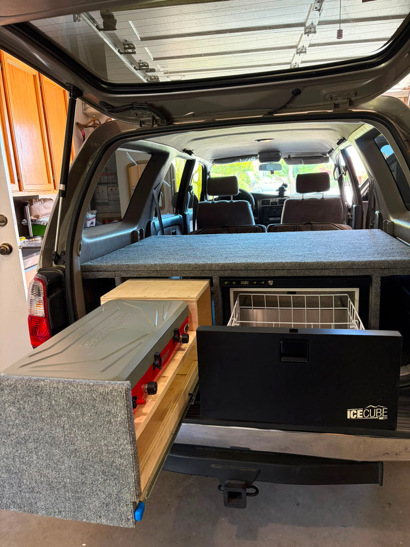 Rear hatch open showing the full camper build: carpeted sleeping platform, slide-out kitchen drawer and Ironman 4x4 IceCube 40L fridge/freezer — photo 2