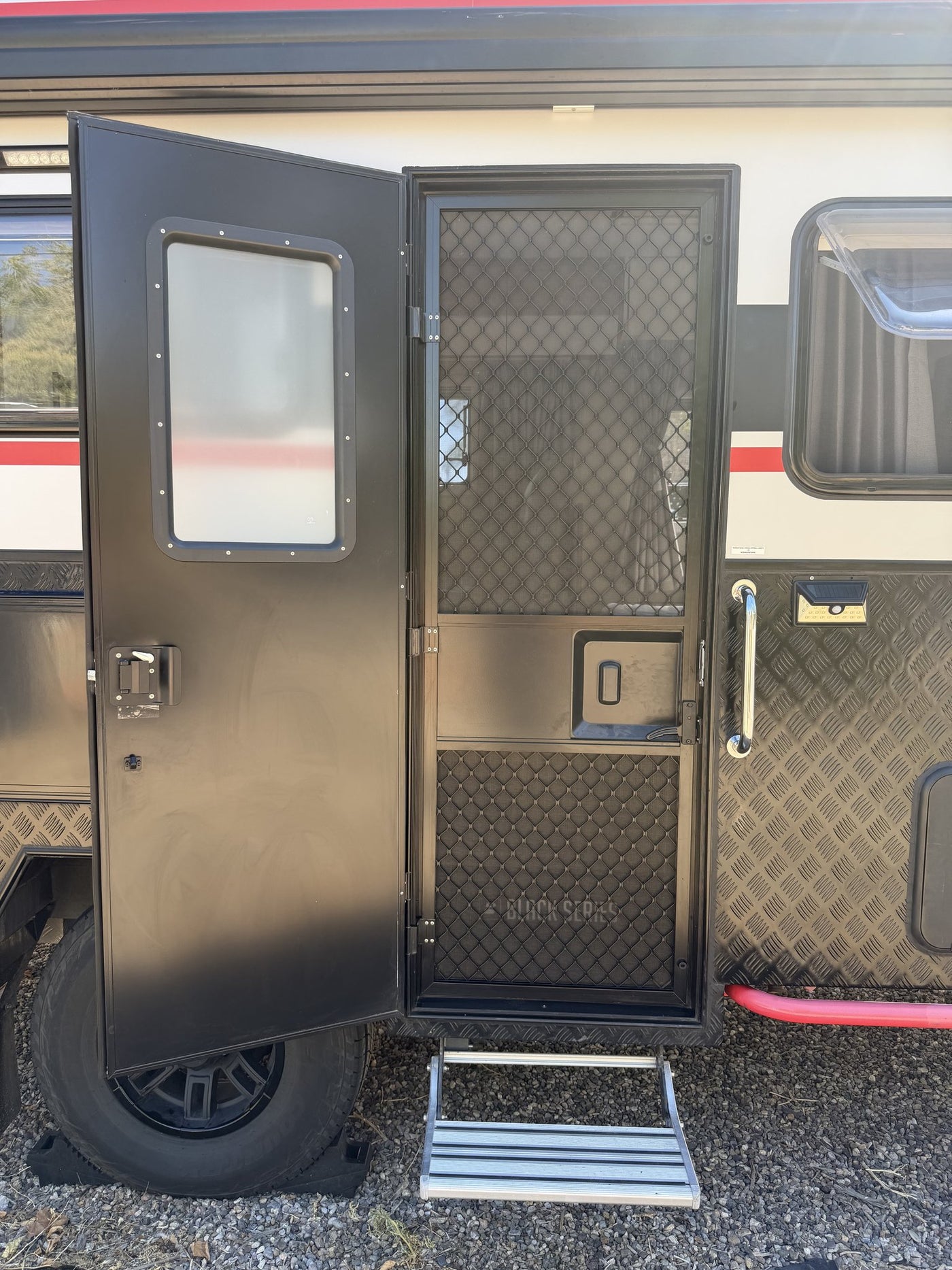 Entry door standing open with the mesh security screen closed, showing the door latch, interior-facing window and the pull-down aluminum entry step over the black alloy wheel — photo 5