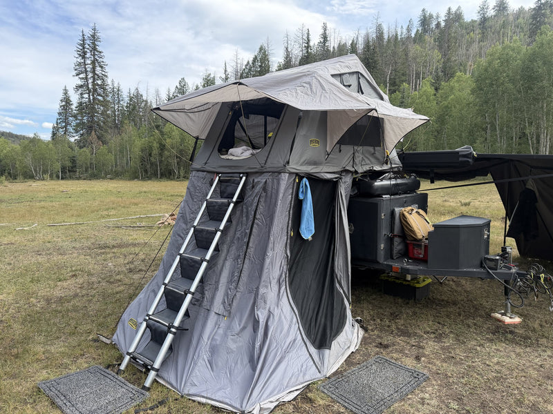 Three-quarter view of the same rooftop tent under blue sky showing the full tent body, the awning-style window covers propped open and the zip-on annex enclosing the ladder to form a private ground room — photo 2