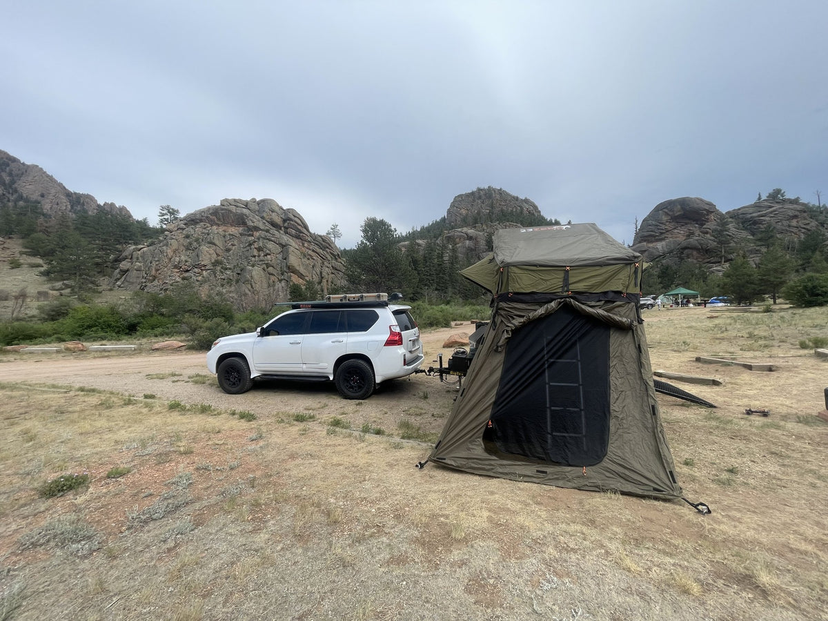 The trailer set up at a Colorado camp with the 23ZERO Walkabout rooftop tent deployed on the elevated platform and the matching annex zipped in around the ladder, hitched behind a white Lexus GX with granite outcrops in the background — photo 2