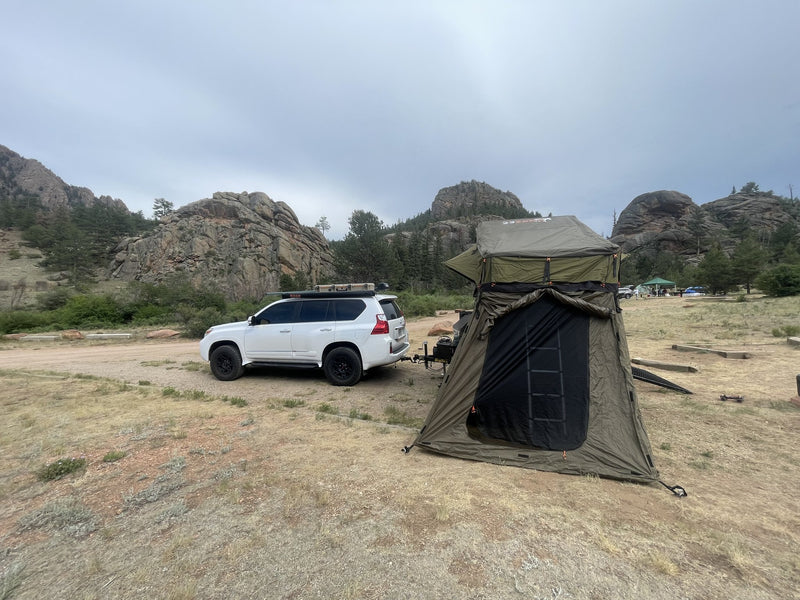 The trailer set up at a Colorado camp with the 23ZERO Walkabout rooftop tent deployed on the elevated platform and the matching annex zipped in around the ladder, hitched behind a white Lexus GX with granite outcrops in the background — photo 2