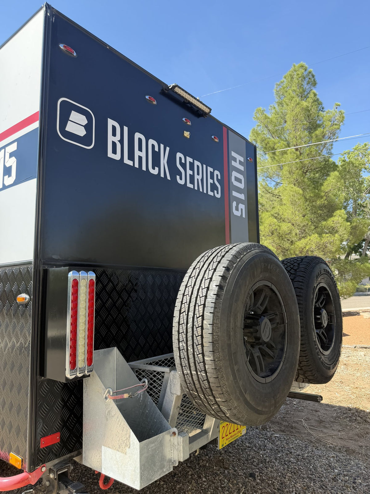 Rear three-quarter view of the trailer showing the Black Series HQ15 tailgate graphics, LED tail light bars, galvanized jerry-can and battery box on the rear bumper, and the two rear alloy spare wheels fitted with new rubber — photo 3