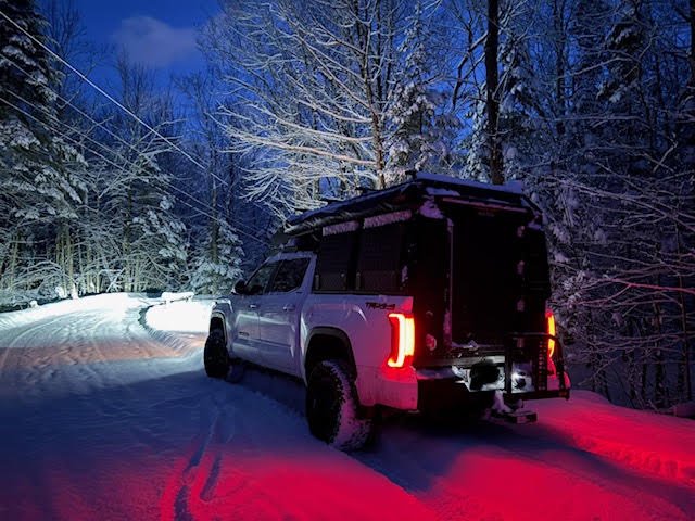 The rig on a snow-covered forest road at dusk with the tail lights lit, camper closed, snow-laden trees on both sides — photo 12