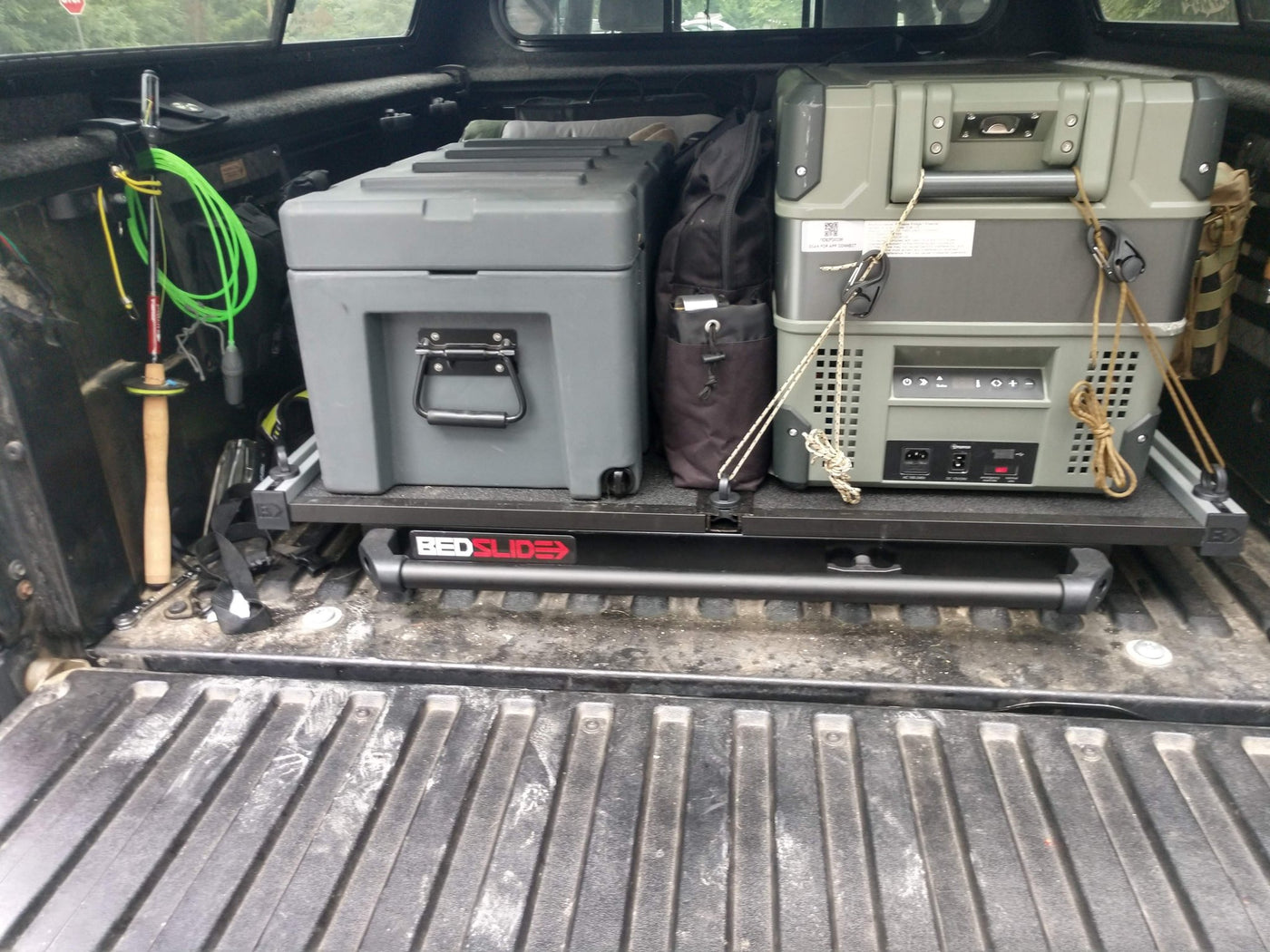 The included BedSlide pulled out over the open tailgate of the Tacoma, its BEDSLIDE-branded front rail visible, loaded with a hard-shell storage case and a portable fridge/freezer lashed down with rope, and gear hung along the bed walls — photo 2
