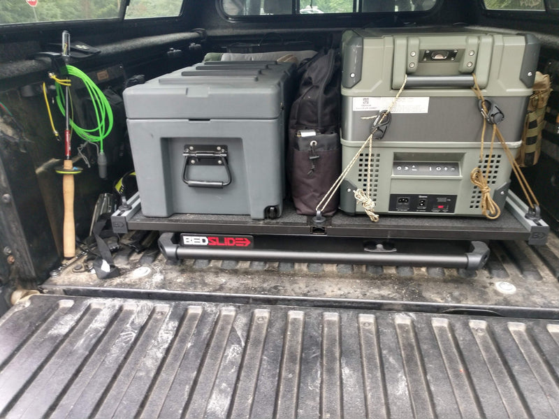 The included BedSlide pulled out over the open tailgate of the Tacoma, its BEDSLIDE-branded front rail visible, loaded with a hard-shell storage case and a portable fridge/freezer lashed down with rope, and gear hung along the bed walls — photo 2