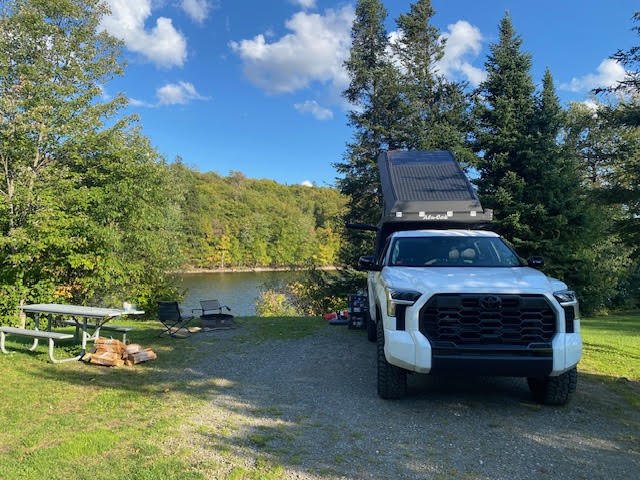 Lakeside campsite in autumn with the Tundra parked beside a picnic table and fire ring, camper roof raised and camp chairs set out by the water — photo 11