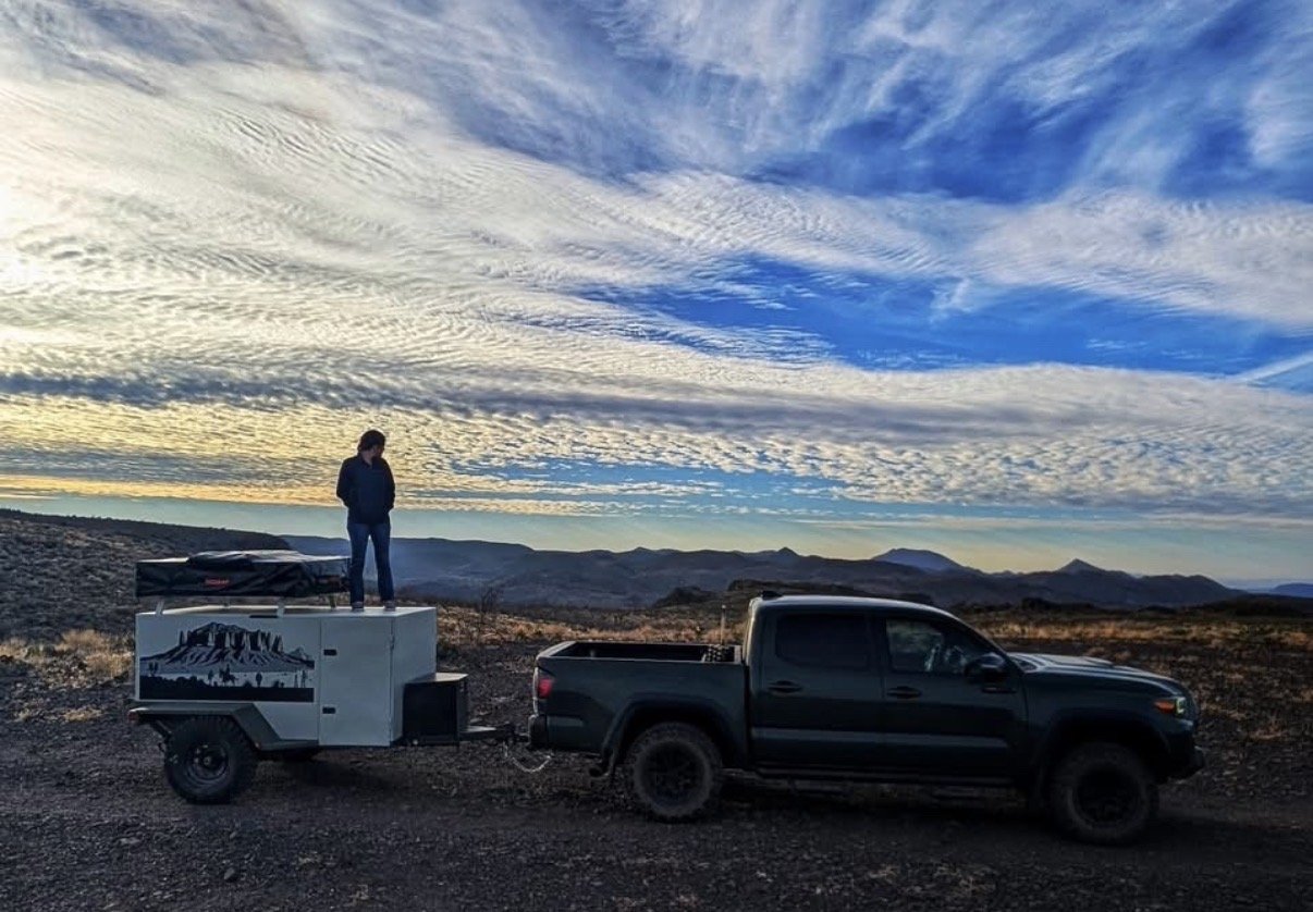 The trailer hitched behind a Toyota Tacoma at a high desert overlook at sunset, a person standing on the trailer roof beside the packed rooftop tent, mountains and streaked clouds in the distance — photo 6