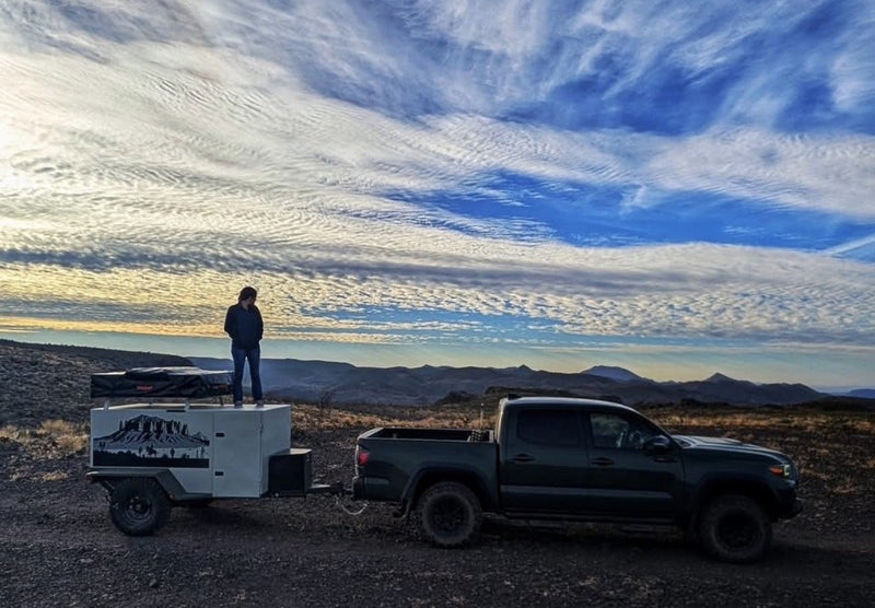 The trailer hitched behind a Toyota Tacoma at a high desert overlook at sunset, a person standing on the trailer roof beside the packed rooftop tent, mountains and streaked clouds in the distance — photo 6
