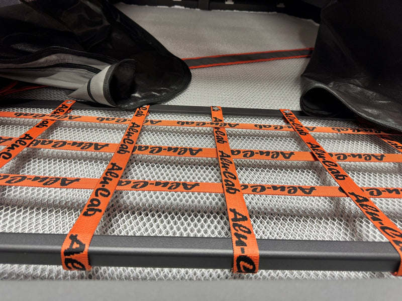 Close view of the rooftop tent's sleeping platform with the white breathable mesh mattress base and the orange Alu-Cab branded webbing straps laid across it — photo 6