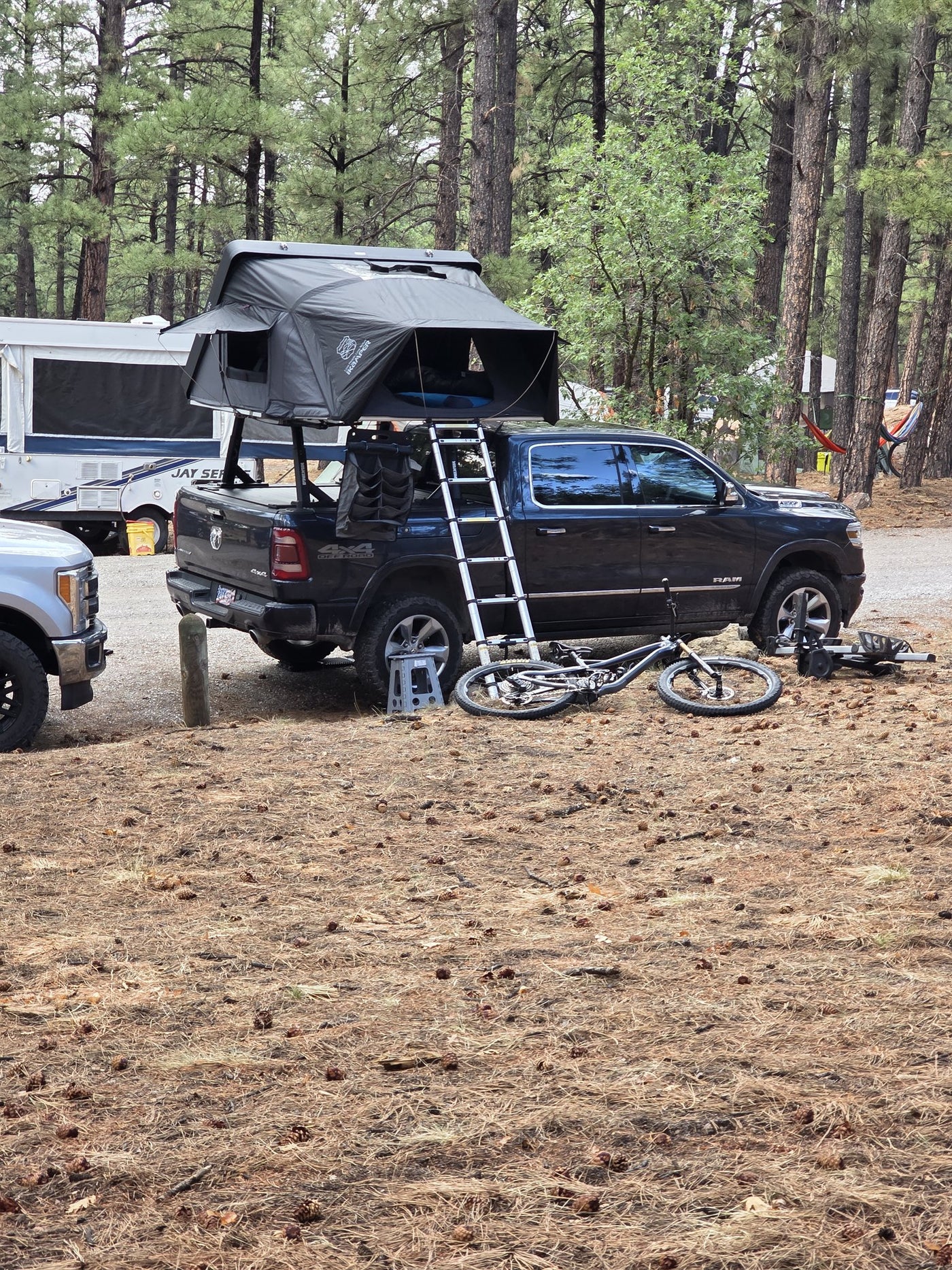 iKamper DLX rooftop tent set up at a wooded campsite, rear three-quarter view with the ladder extended — photo 3