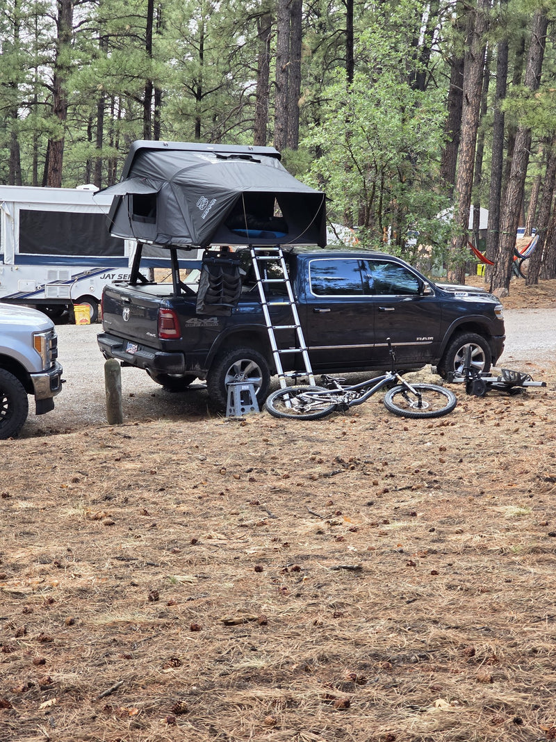 iKamper DLX rooftop tent set up at a wooded campsite, rear three-quarter view with the ladder extended — photo 3
