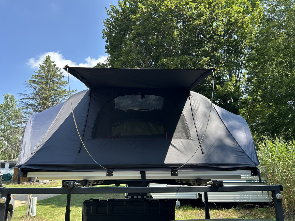 iKamper X-Cover 2.0 rooftop tent opened on a vehicle rack against a blue sky, straight-on front view showing the full grey and black tent body, the extended rainfly canopy over the door and the large mesh window