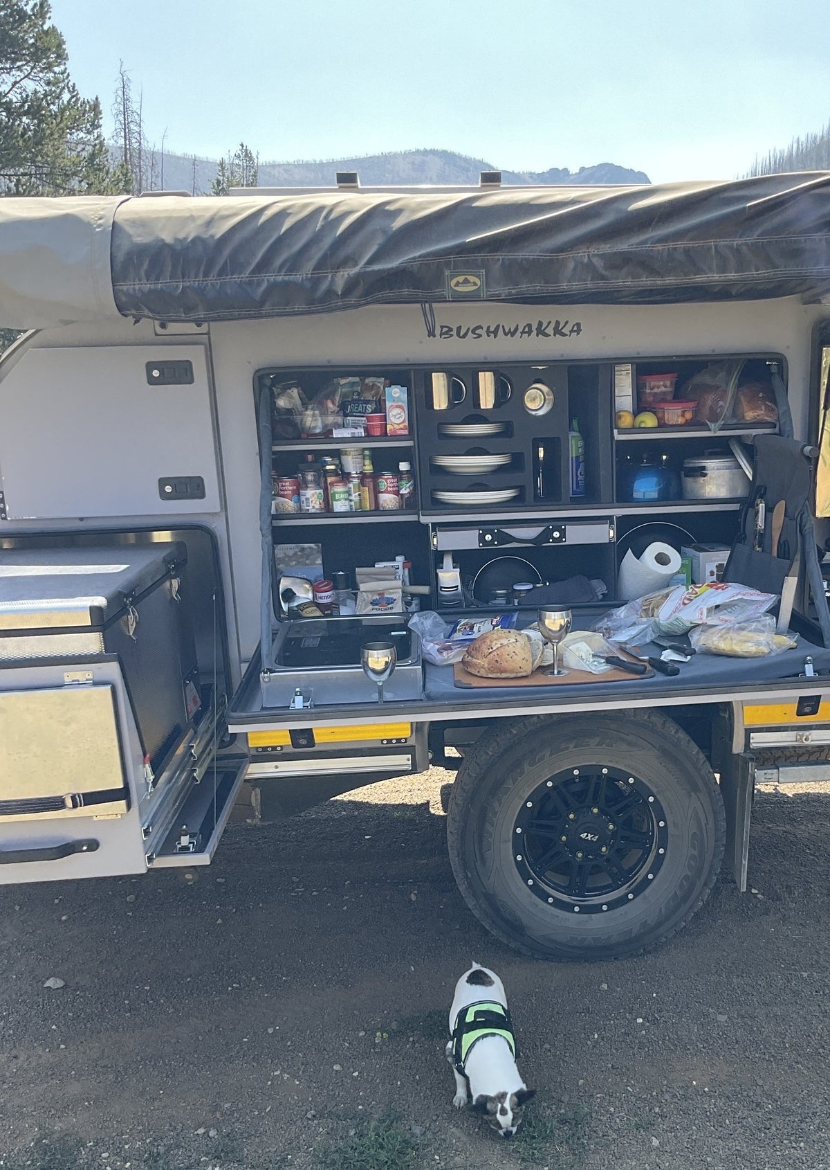 The Bushwakka slide-out camp kitchen fully deployed under the side awning: stocked pantry shelving, plate and mug racks, two-burner stove, fold-down prep counter in use, drawer units and the spare wheel on the black alloy rim — photo 3