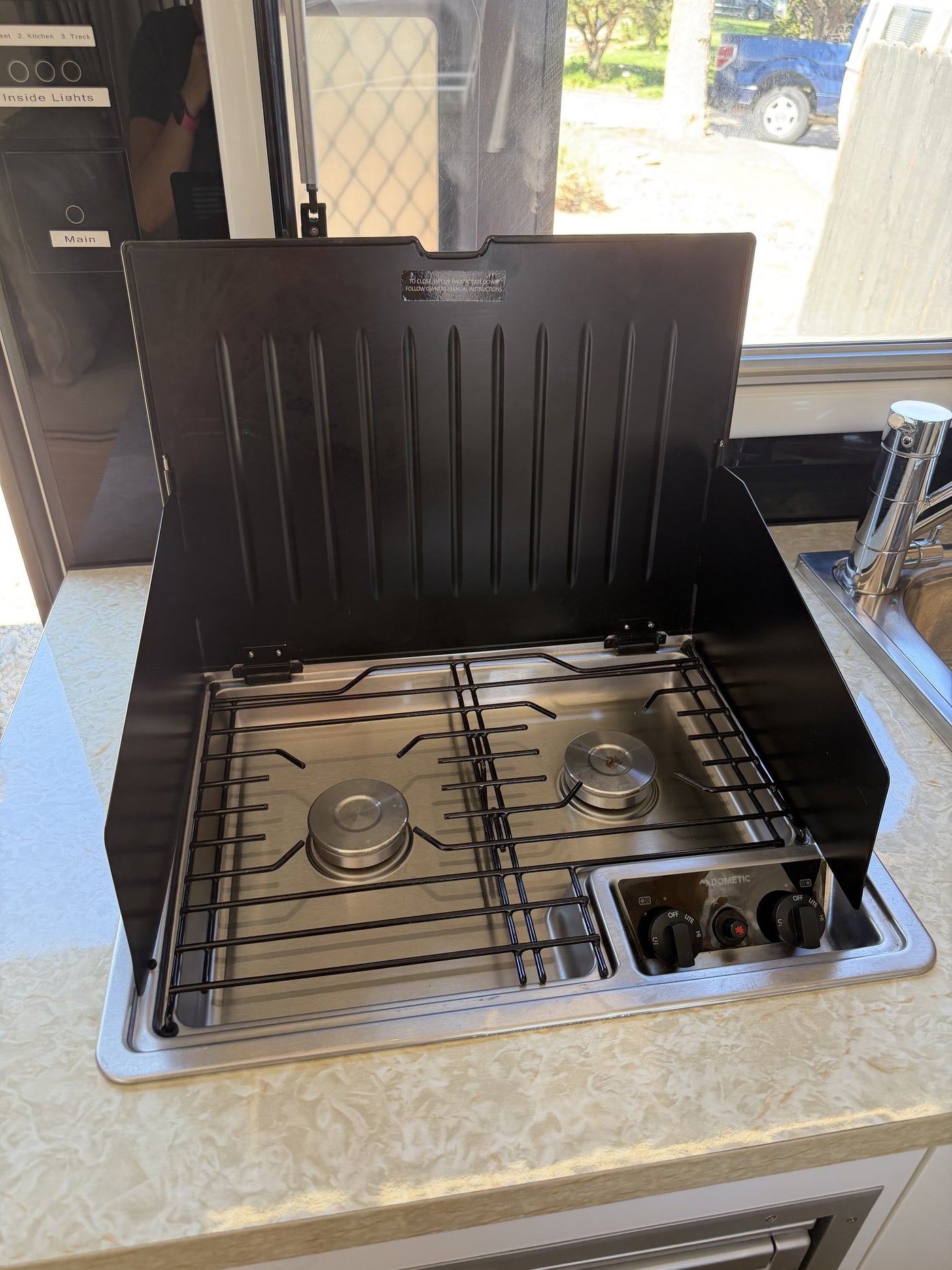 Dometic two-burner gas cooktop with the glass lid raised, showing the burner grates, control knobs and the adjacent sink on the stone-look countertop — photo 11