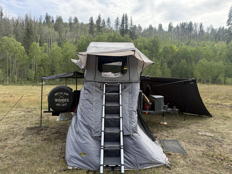 Smittybilt Gen2 softshell rooftop tent set up on a trailer in a mountain meadow, straight-on front view with the rainfly extended, the mesh door open and the telescoping aluminum ladder running down through the grey annex room below — photo 1