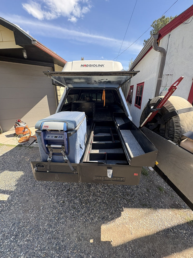 Rear view of the Extendobed system slid out of the truck bed under a canopy and rooftop tent, showing the divided drawer compartments and the strapped ARB fridge — photo 2