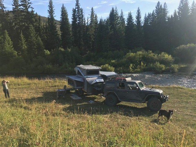 Riverside meadow camp in evening light with the Sundowner's rooftop tent and awning deployed beside the Jeep, spruce timber and a creek behind — photo 4