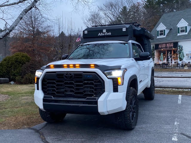 Front view of the Tundra in winter with the Alu-Cab Cabin raised, showing the TRD grille with amber marker lights, AVS Aeroskin hood protector and the roof-rack cargo box above the cab — photo 5