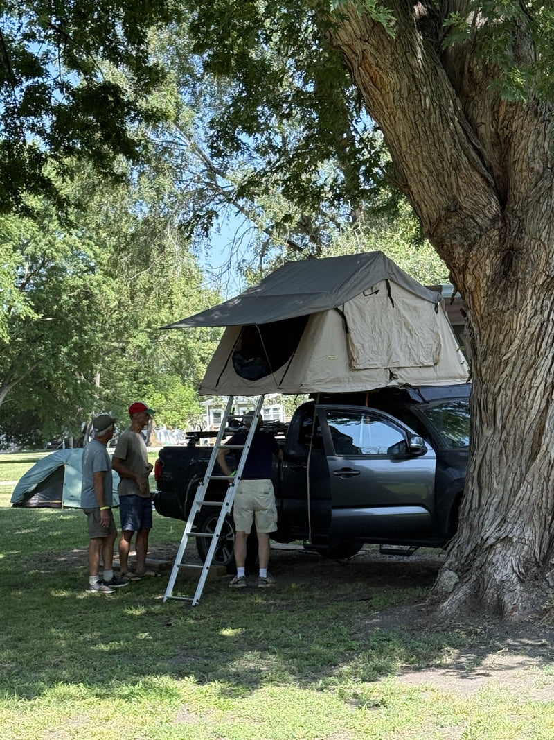 Tent deployed on the truck under a large shade tree, awning extended over the entry and ladder angled to the grass — photo 4