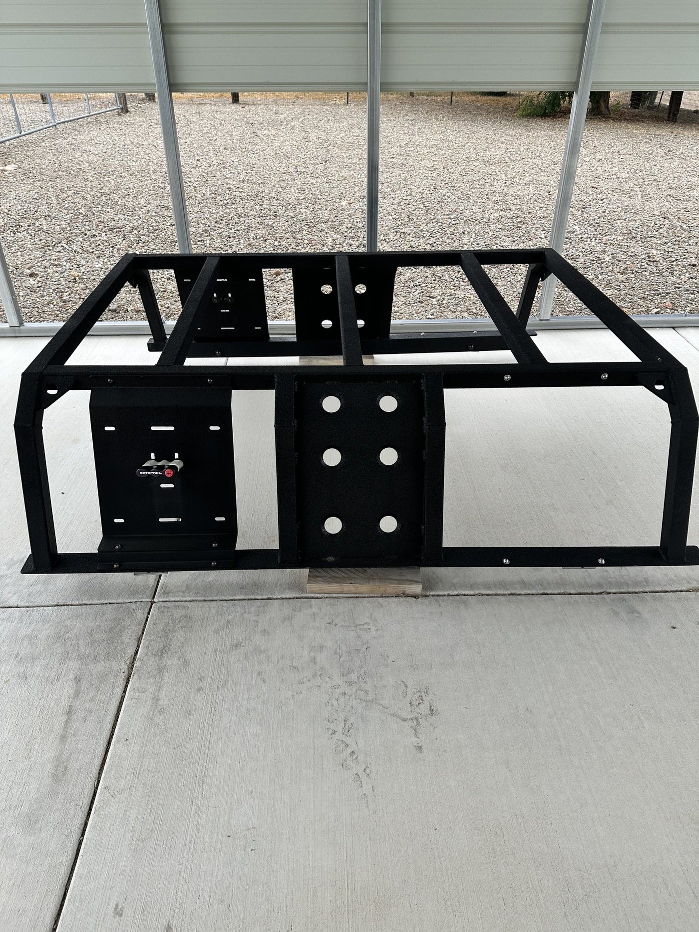 Black Nuthouse Nutzo Tech 1 aluminum expedition bed rack standing on a concrete pad under a carport, front view showing the full welded frame, two universal mounting plates and a Rotopax locking mount — photo 1