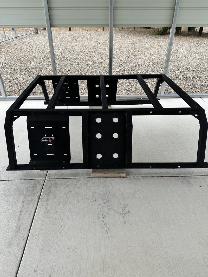 Black Nuthouse Nutzo Tech 1 aluminum expedition bed rack standing on a concrete pad under a carport, front view showing the full welded frame, two universal mounting plates and a Rotopax locking mount — photo 1