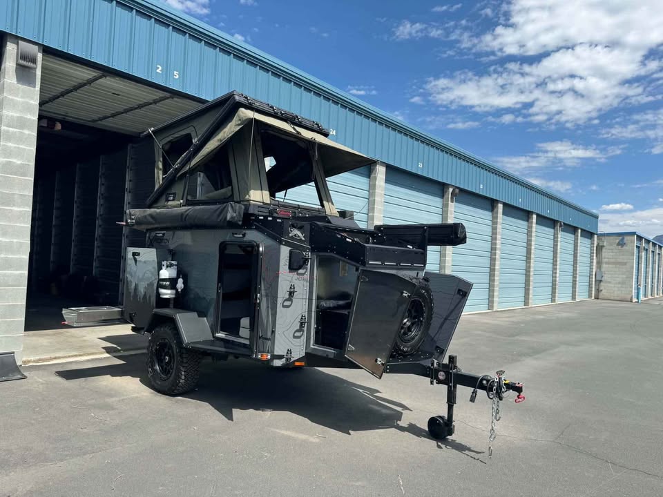 2025 XPedition Voyager Sky XE overland trailer parked outside a storage unit in the sun, three-quarter view from the tongue, with the Bush Company TX27 MAX hardshell rooftop tent popped open, the side galley hatch raised, spare tire on the rear swing-out and off-road tires on black wheels — photo 1