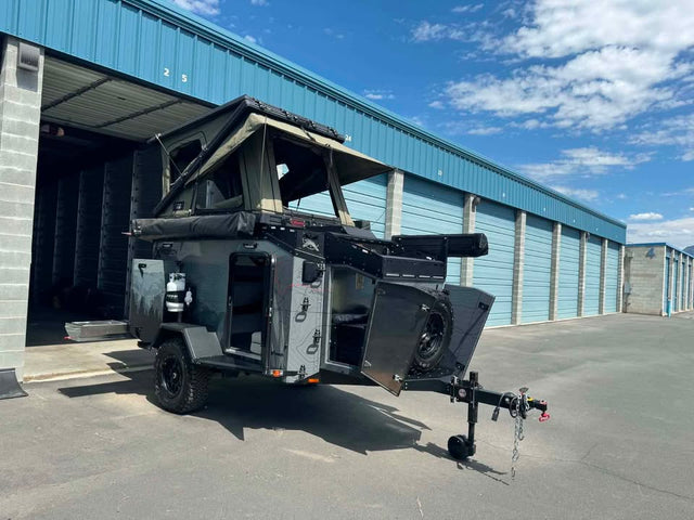 2025 XPedition Voyager Sky XE overland trailer parked outside a storage unit in the sun, three-quarter view from the tongue, with the Bush Company TX27 MAX hardshell rooftop tent popped open, the side galley hatch raised, spare tire on the rear swing-out and off-road tires on black wheels