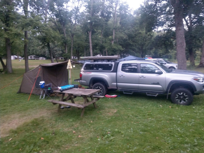 Oztent RV-4 set up at a shaded campsite under overcast light, showing the side panel, mesh window and extended awning over a picnic table — photo 2