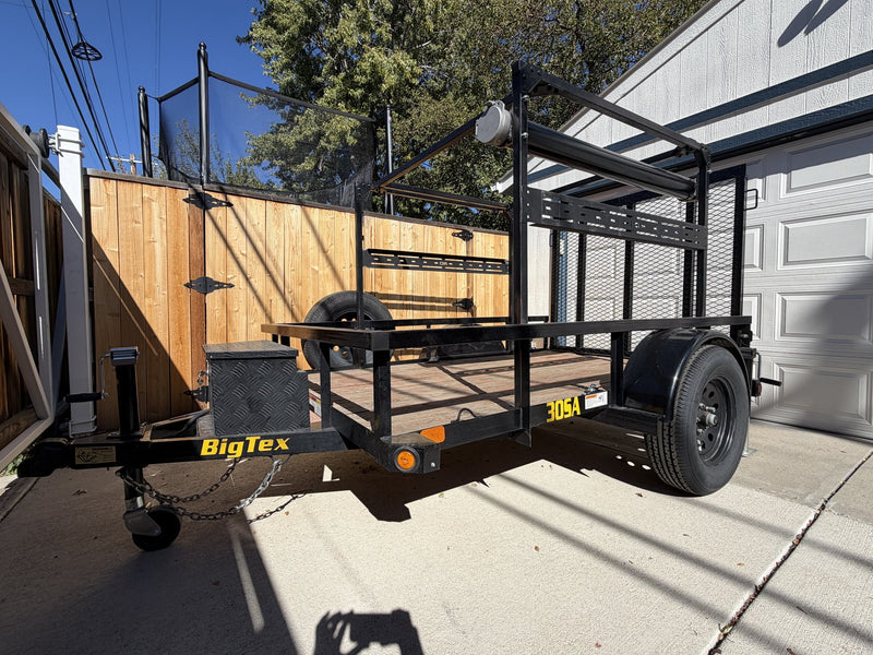 Front three-quarter view from the driver side showing the A-frame tongue with coupler and safety chains, the diamond-plate aluminum storage box on the tongue, the spare tire carried on the deck and the full height of the overhead rack frame — photo 2