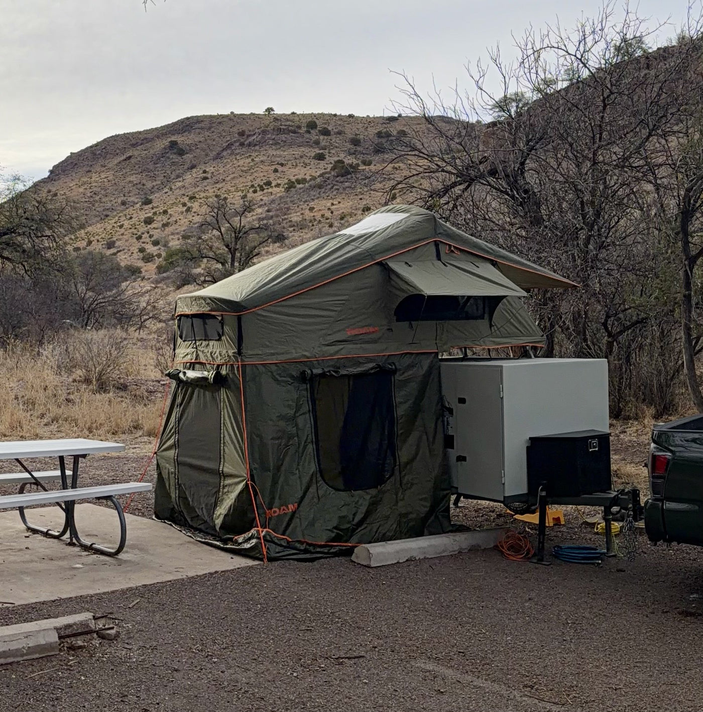 The trailer set up at a desert campsite with the Roam Vagabond rooftop tent deployed and its full annex room zipped on to the ground, hitched beside a picnic table with hills behind — photo 2