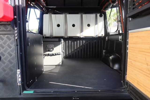 Inside the camper looking forward from the tailgate: carpeted Goose Gear floor panel, 16-gallon fresh water tanks across the bulkhead, aluminium storage cases and the checker-plate and wood interior panels — photo 8
