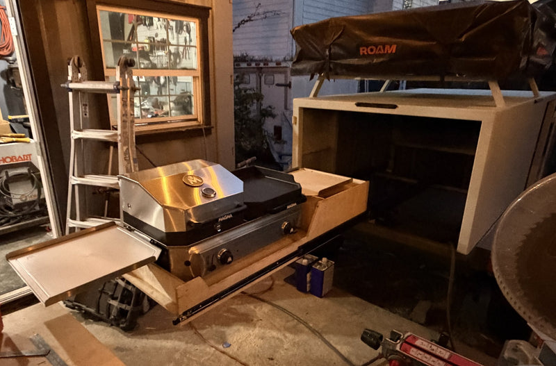 Evening shop view of the driver-side rear slide-out drawer extended with the Magma Crossover double burner mounted and its side prep shelf folded out, rear hatch open and the Roam rooftop tent on the rack — photo 7
