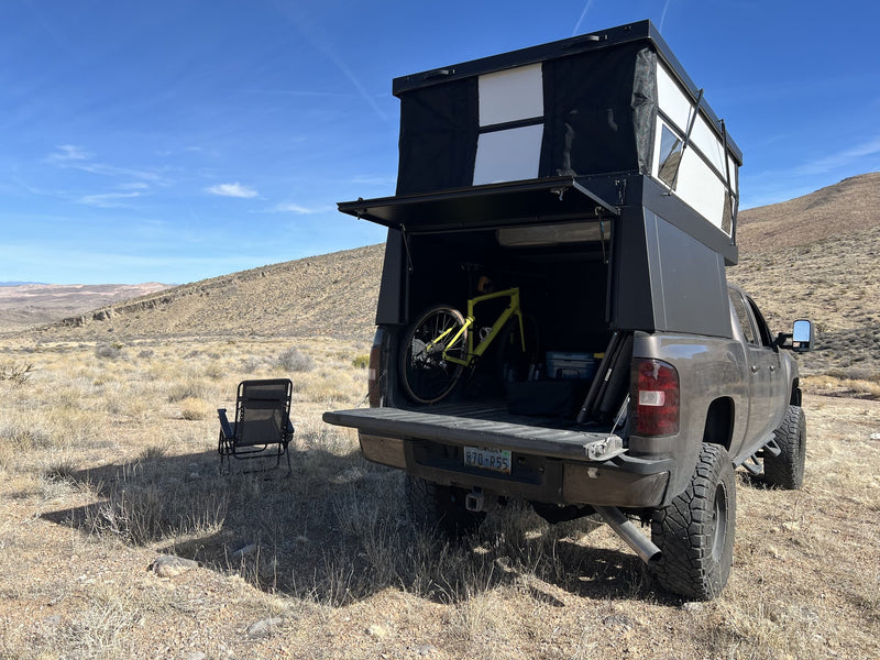 Rear view of the Oru Designs Bruin with the roof raised and the rear hatch lifted, showing the sleeping platform above and open truck-bed storage below with a bike loaded inside — photo 3