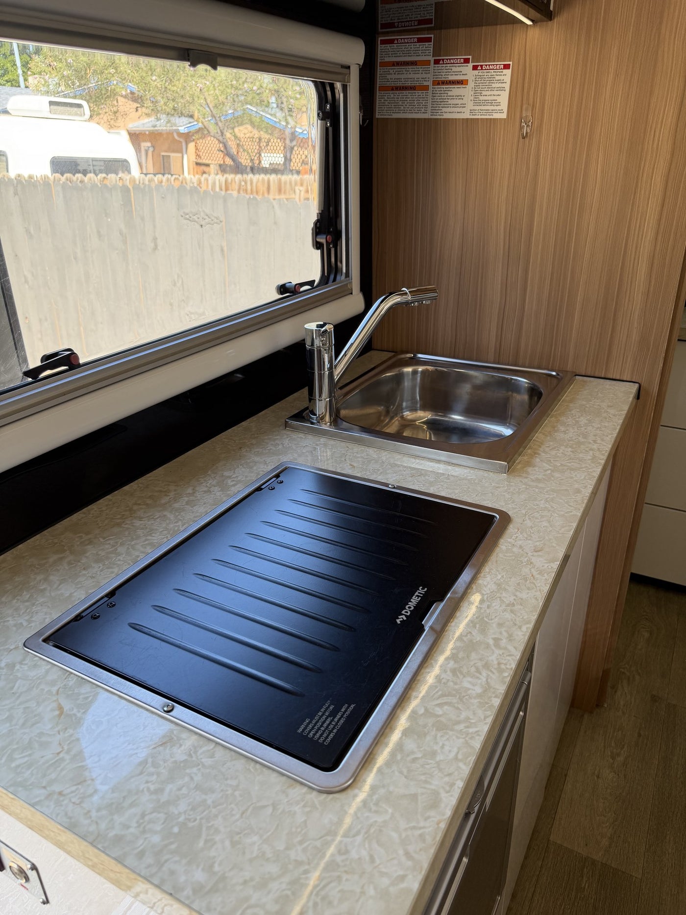 Galley counter in detail: stainless steel sink with pull-out faucet beside the covered Dometic cooktop, opening window over the bench, and the drawer stack and appliance bay below — photo 10