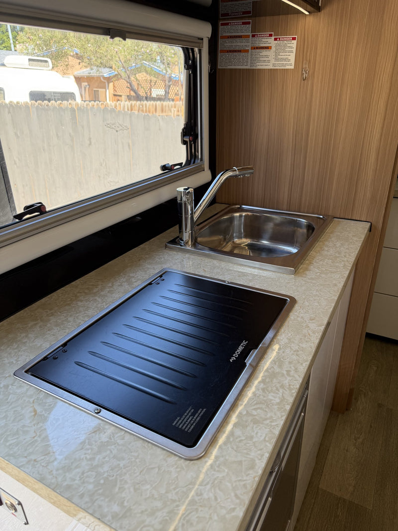 Galley counter in detail: stainless steel sink with pull-out faucet beside the covered Dometic cooktop, opening window over the bench, and the drawer stack and appliance bay below — photo 10