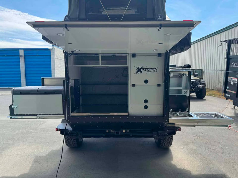 Rear view of the XPedition Voyager Sky XE with the galley hatch raised, showing the XPEDITION TRAILERS logo, open pantry shelving, the slide-out stainless kitchen with two-burner stove on the right and a slide-out cushioned bench on the left — photo 3